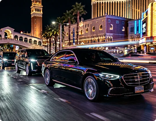 Two black luxury vehicles drive down a brightly lit city street at night, surrounded by neon lights and blurred motion. The scene captures a lively urban setting with palm trees, illuminated buildings, and a sense of speed and sophistication.